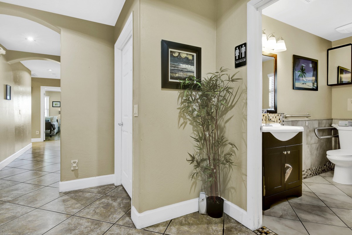 A hallway with tile flooring, featuring a potted plant and artwork on the wall, leading to a bathroom with a mirror and vanity.