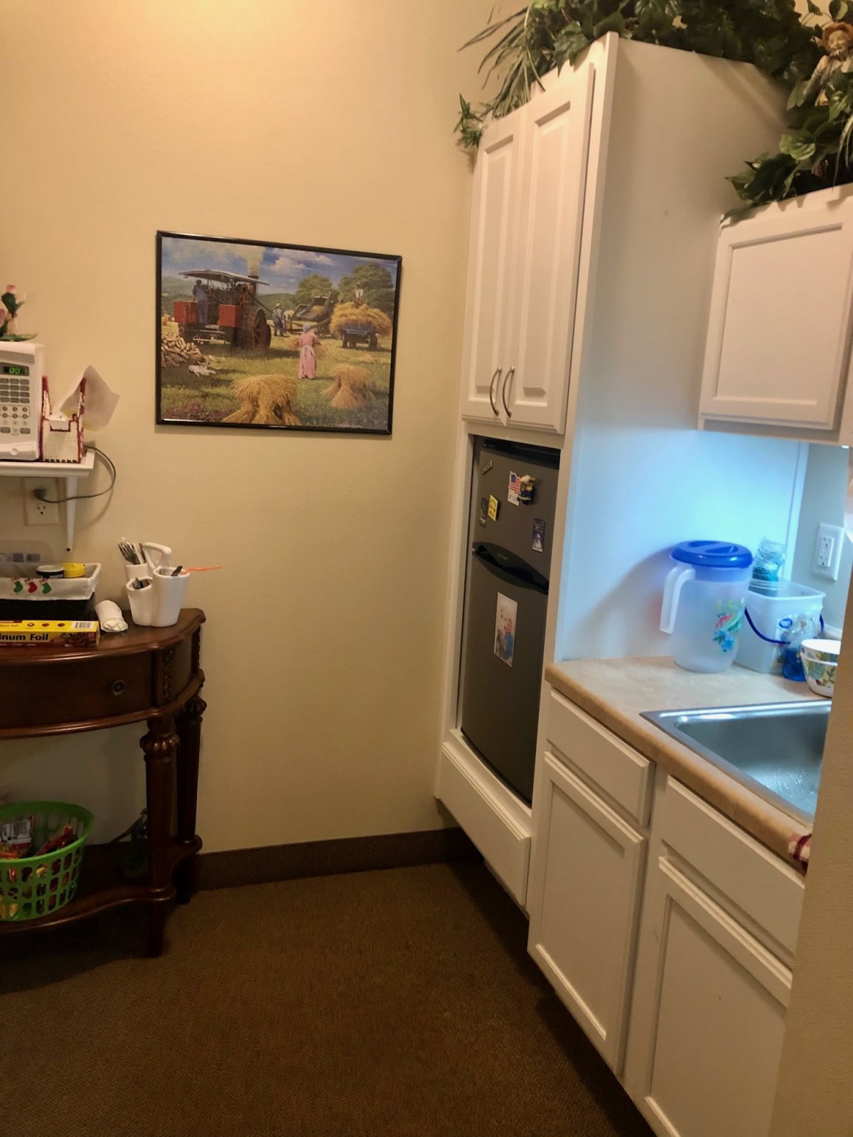 A small kitchen area with cabinets, a sink, microwave, and a painting on the wall.