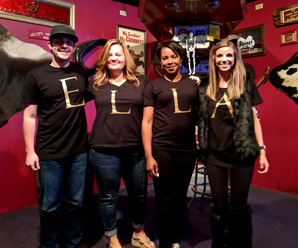 A group of four people wearing matching t-shirts with letters stand together at an indoor event.