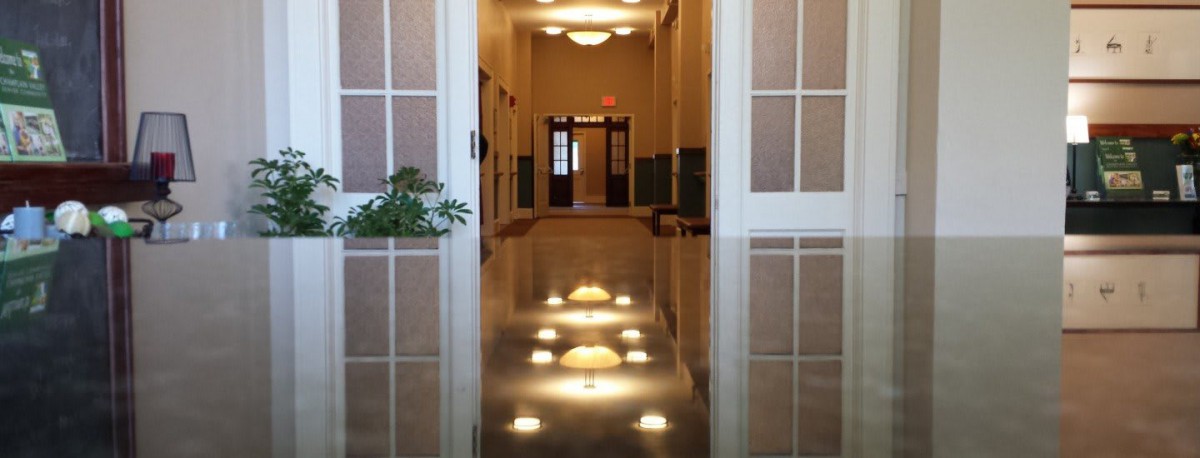 A polished hallway with reflections of ceiling lights on the shiny floor, leading to a set of double doors.