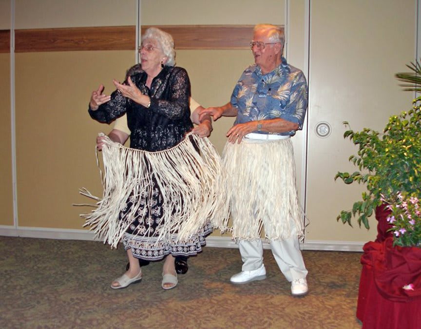 An elderly couple dressed in hula skirts and a Hawaiian shirt enjoying a dance indoors.
