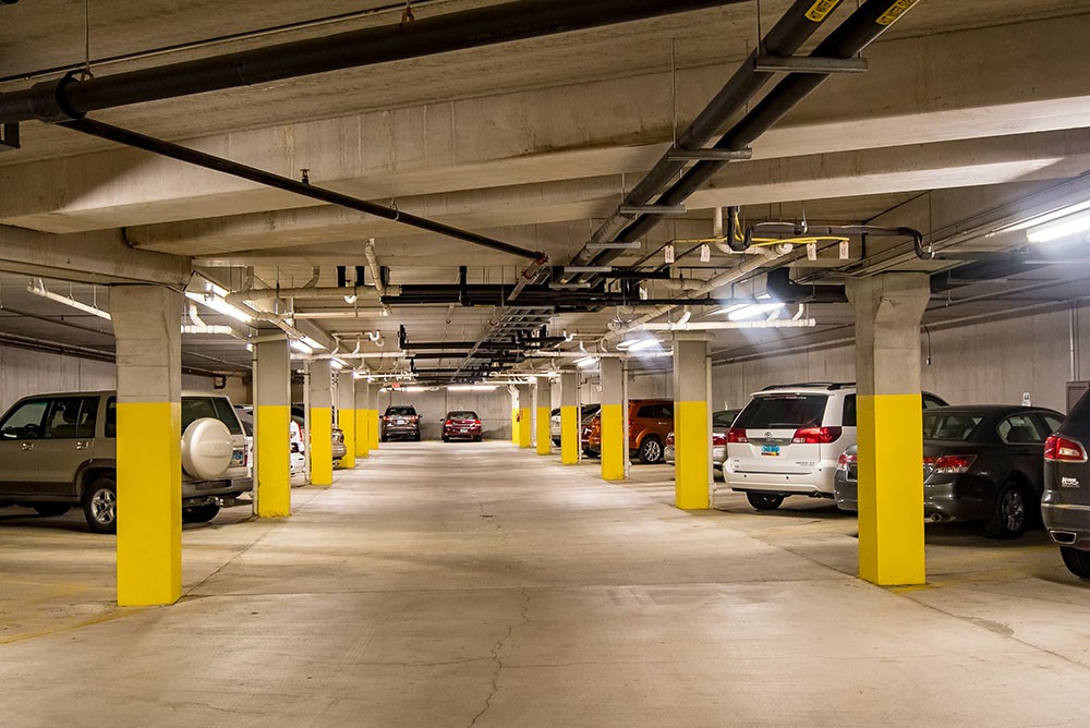 An underground parking garage with several cars parked alongside concrete pillars under fluorescent lights.
