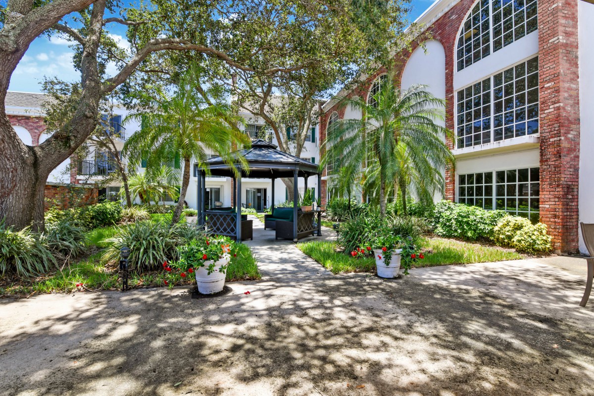 A peaceful garden with a gazebo surrounded by palm trees and greenery in front of a large building.