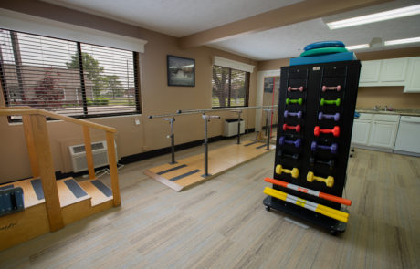 A well-equipped physical therapy room featuring balance bars and a rack of colorful dumbbells.