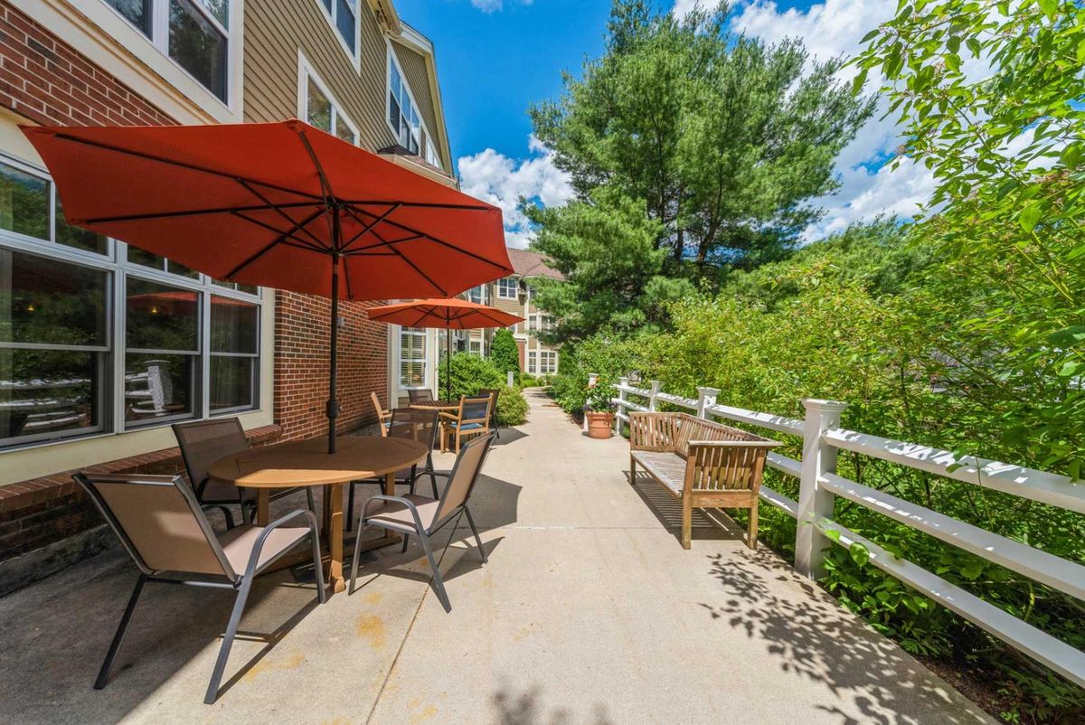 A sunny outdoor patio area with tables, chairs, and red umbrellas surrounded by greenery.