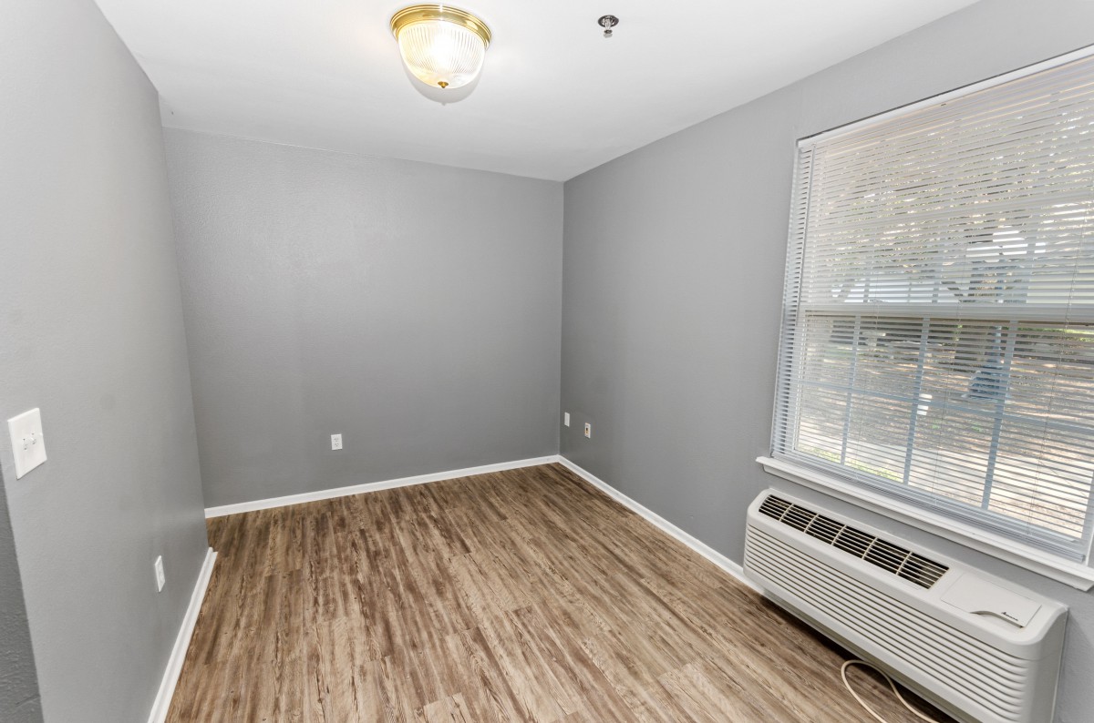 An empty room with gray walls, wood flooring, a ceiling light, a window, and an air conditioning unit.