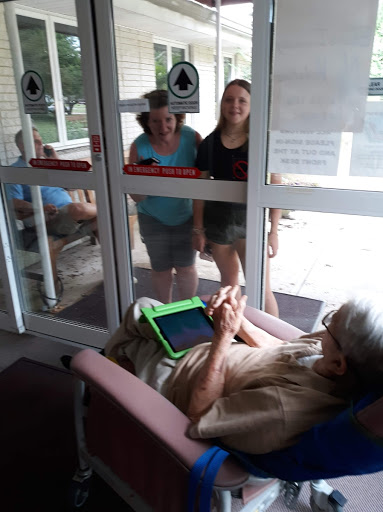 An elderly man sits indoors using a tablet while two girls watch him from outside the glass door.