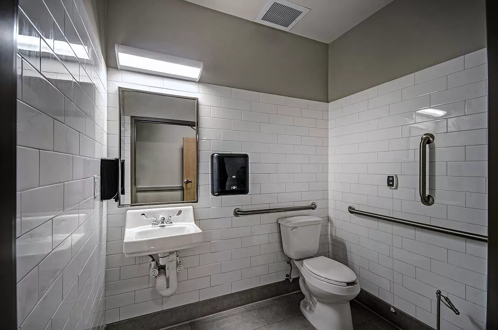 A clean and accessible bathroom with white tiled walls, featuring a toilet, sink, mirror, and grab bars.