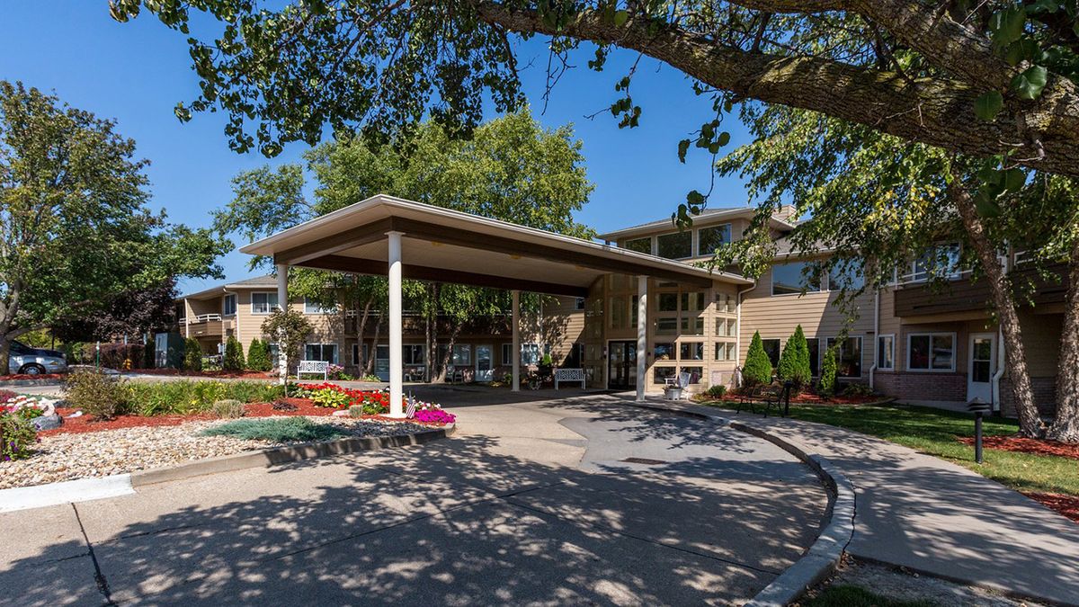 A covered entrance to a building surrounded by trees and colorful flowers on a sunny day.