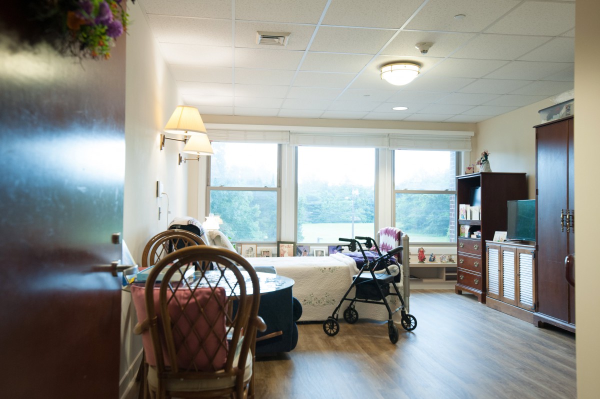 A well-lit hospital room with a bed, wheelchair, and large windows providing a view.