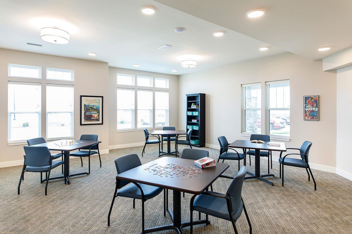 A spacious and well-lit room with multiple tables and chairs, featuring large windows and a bookshelf.