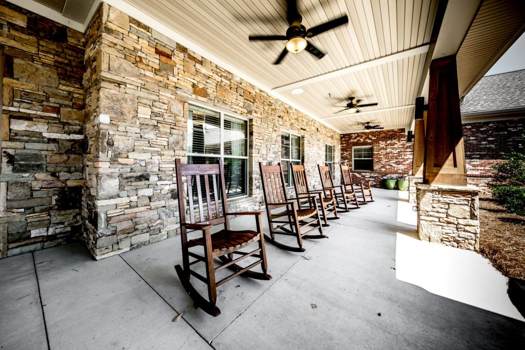 A cozy porch featuring a row of wooden rocking chairs and ceiling fans beneath a stone facade.