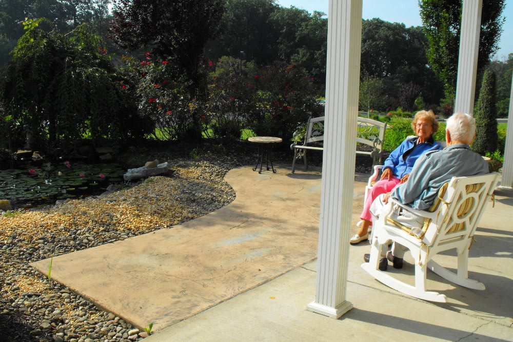 An elderly person sits on a patio bench on a sunny day, overlooking a garden path.