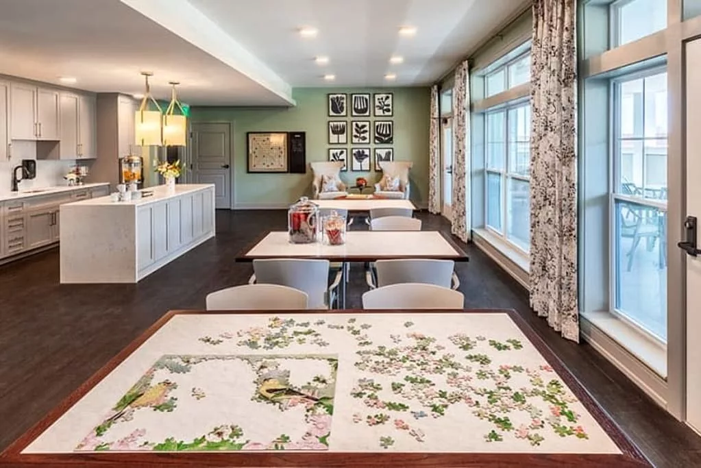 A well-lit room with tables set up for a puzzle activity, featuring decorative art on the walls and large windows.