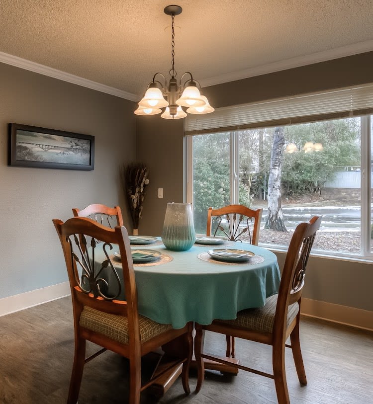 A cozy dining room with a round table set for four, illuminated by a hanging chandelier and bathed in natural light from a large window.