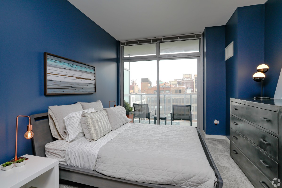 A modern bedroom with blue walls, featuring a neatly made bed and large windows leading to a balcony.
