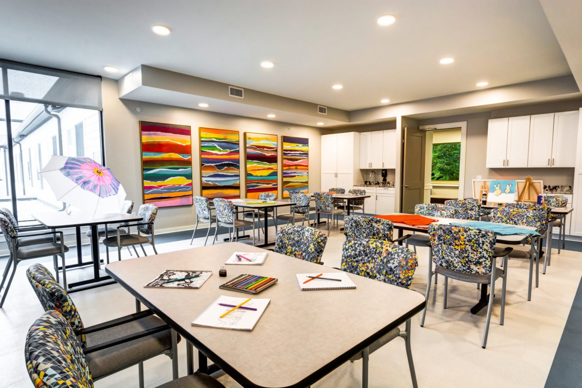 A bright and colorful art gallery space with tables, chairs, and vibrant paintings on the walls.