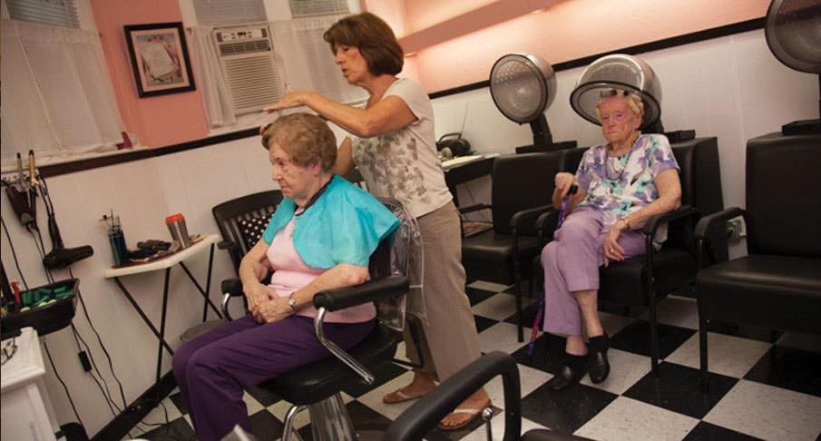 A hair stylist working on an elderly woman's hair while another woman sits under a hair dryer in a salon.