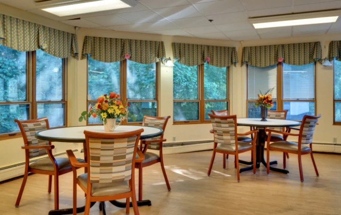 A cozy dining room with tables and chairs, decorated with flowers and surrounded by large windows with striped curtains.