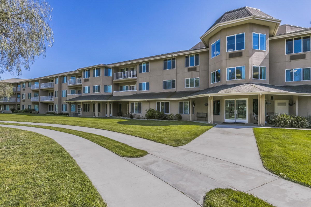 A three-story residential apartment building with a well-maintained lawn and pathways on a sunny day.