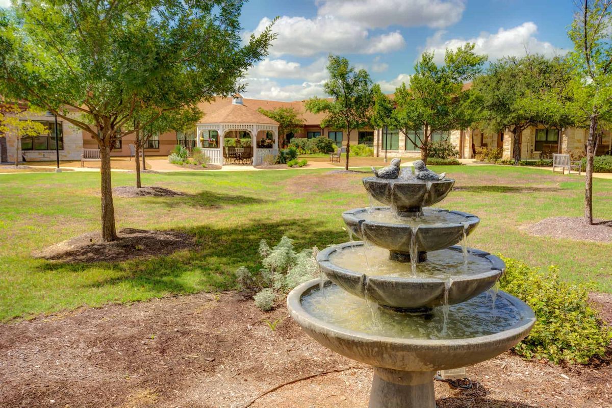 A serene garden features a tiered fountain and a gazebo surrounded by trees under a blue sky.