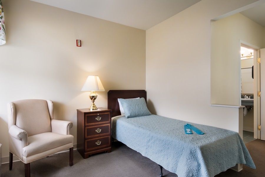 A minimalist bedroom featuring a single bed, a nightstand with a lamp, and an upholstered chair.