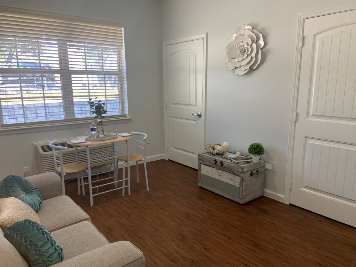 A cozy living room with minimalist decor, featuring a small dining table, wooden floor, and ample natural light from a window with blinds.