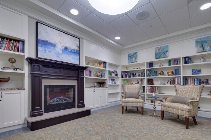 A cozy reading nook with a fireplace and bookshelves, featuring comfortable chairs and abstract artwork.