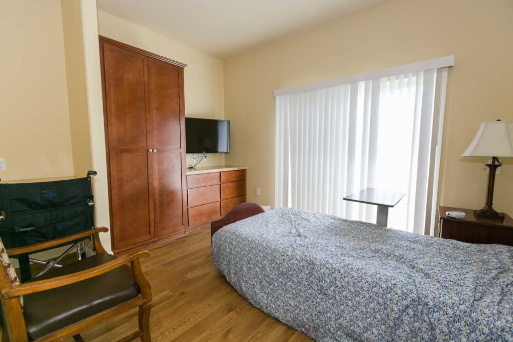 A modern bedroom with minimalist decor, featuring a single bed, wooden floors, a TV on a dresser, and large window blinds.