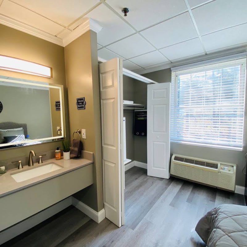 A modern bedroom with an adjacent bathroom area featuring a sink and mirror, bright window, and open closet.
