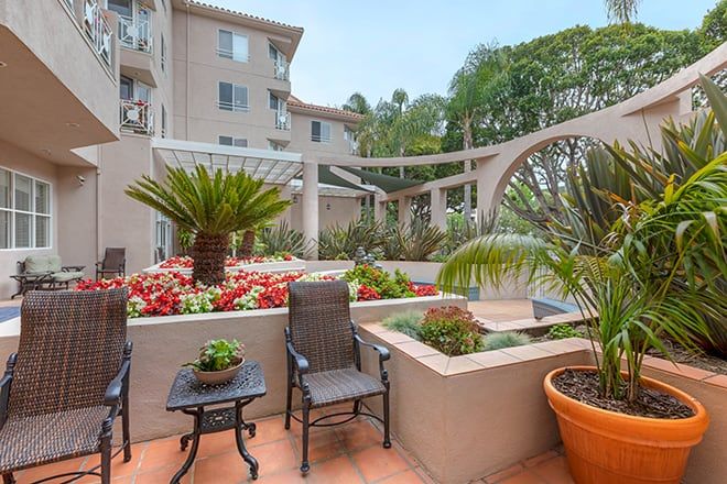 A cozy patio with seating surrounded by lush plants and flowers, adjacent to an apartment building.