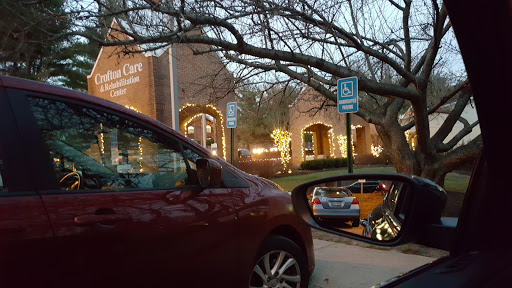 A red car parked in a lot, reflecting on the side mirror, with a brightly lit building and tree branches visible in the evening.