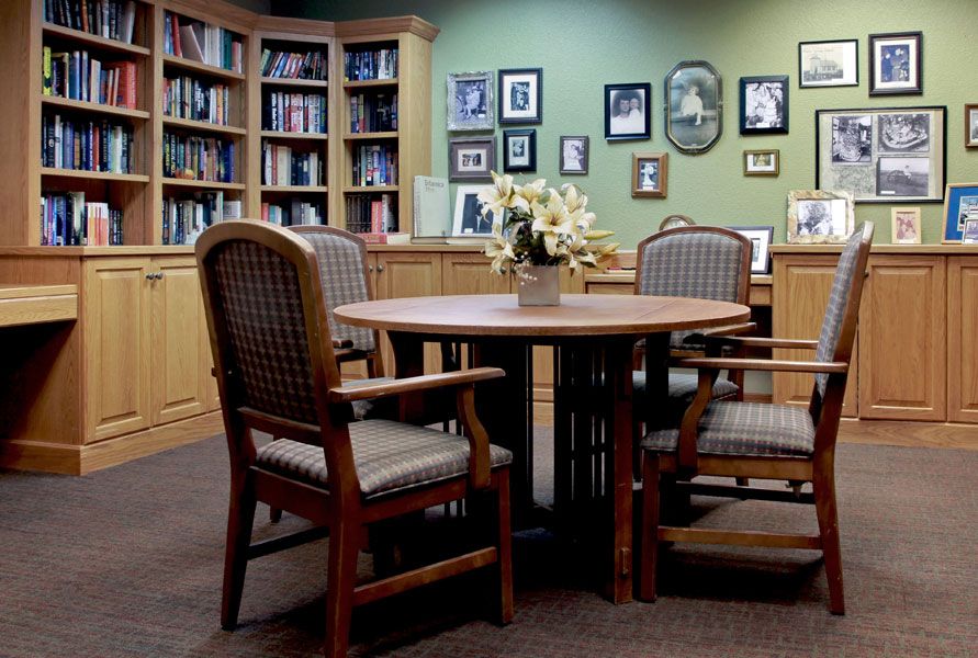 A cozy library meeting area with a round table surrounded by chairs, bookshelves filled with books, and a wall adorned with framed photos.