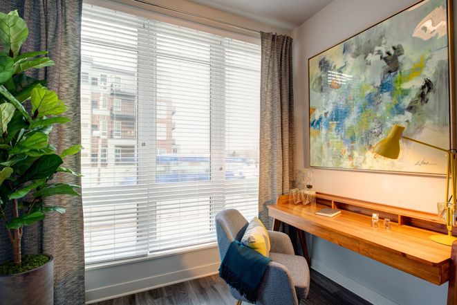 A modern workspace with a wooden desk, comfortable chair, large window with blinds, and abstract artwork on the wall.