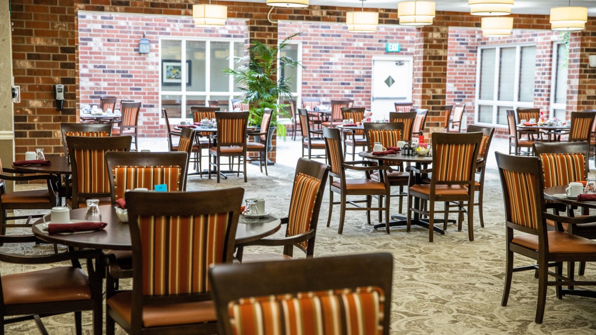 An empty restaurant dining area featuring wooden tables and chairs against a brick wall backdrop.