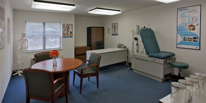 A clean and organized doctor's examination room with a bed, chairs, table, and medical equipment.
