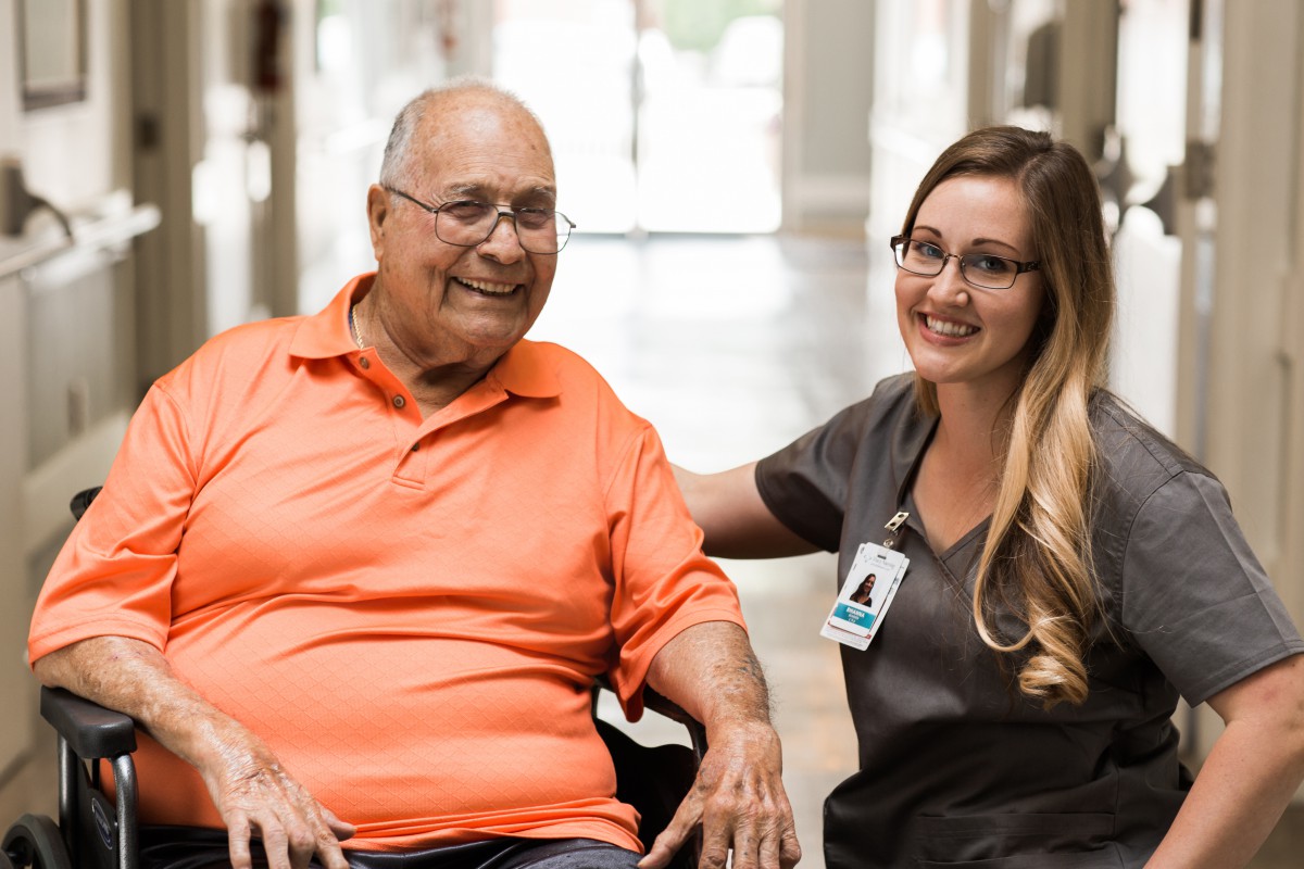 An elderly person in a wheelchair smiling with a caregiver inside a healthcare facility.
