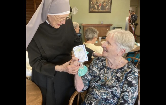 An elderly woman smiles as a nun hands her a card, showcasing joyful interaction in a nursing home setting.