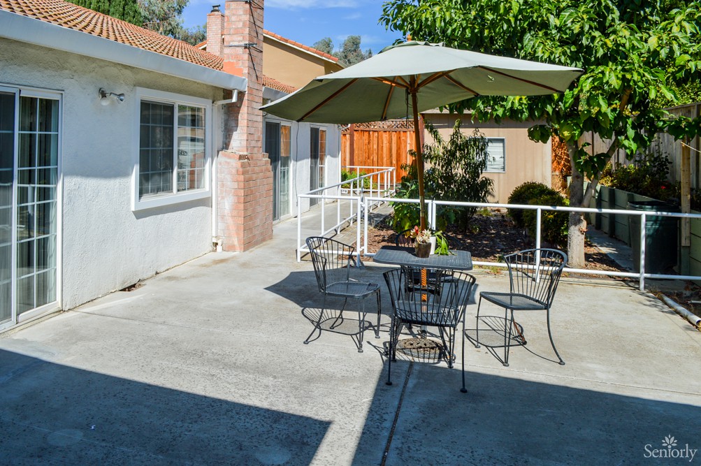 A sunny patio with a table and chairs set under an umbrella, surrounded by greenery.
