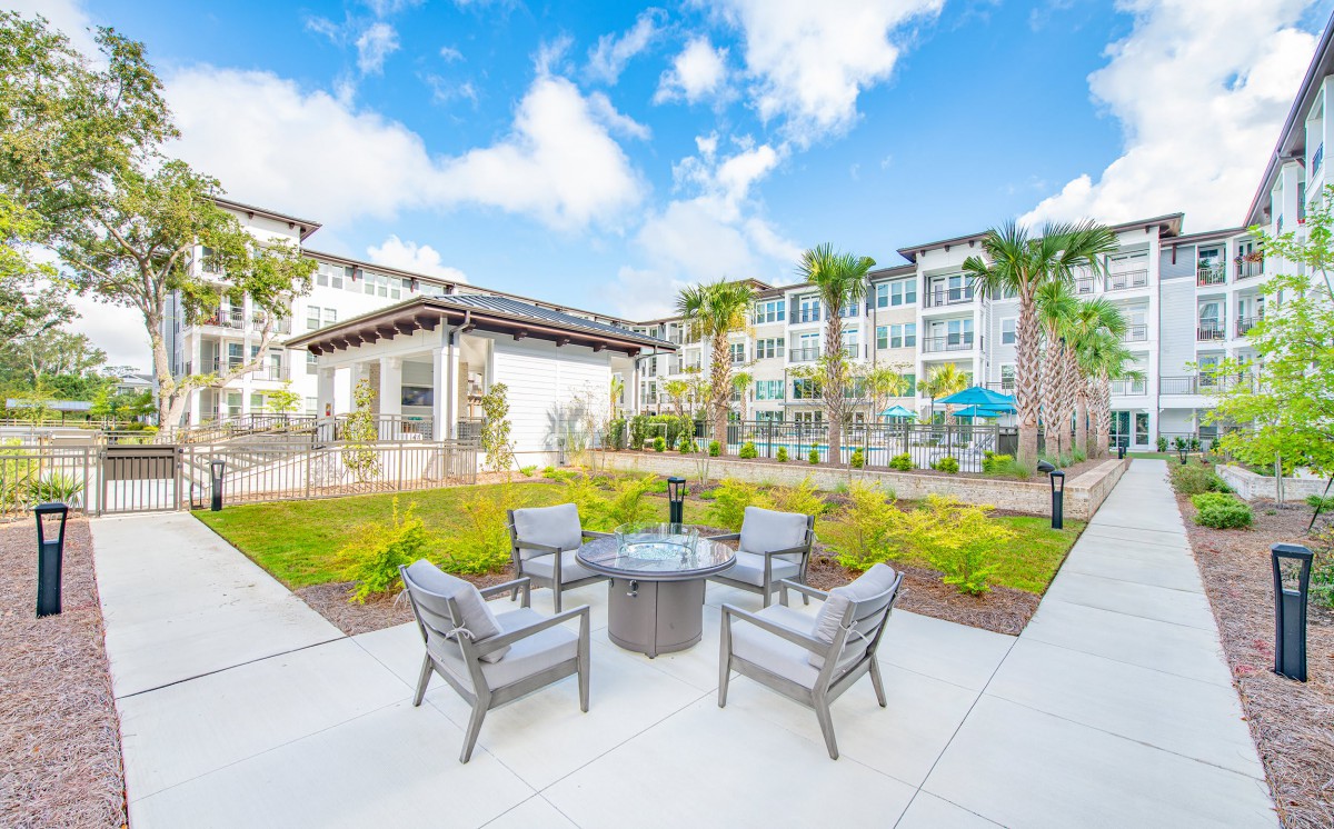 A modern apartment complex featuring an outdoor seating area with chairs and a table, surrounded by well-maintained landscaping under a clear blue sky.