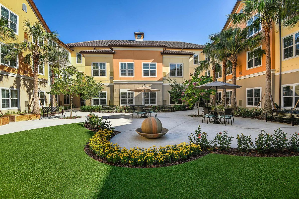A beautifully landscaped courtyard featuring a central fountain, surrounded by a building with outdoor seating and palm trees.