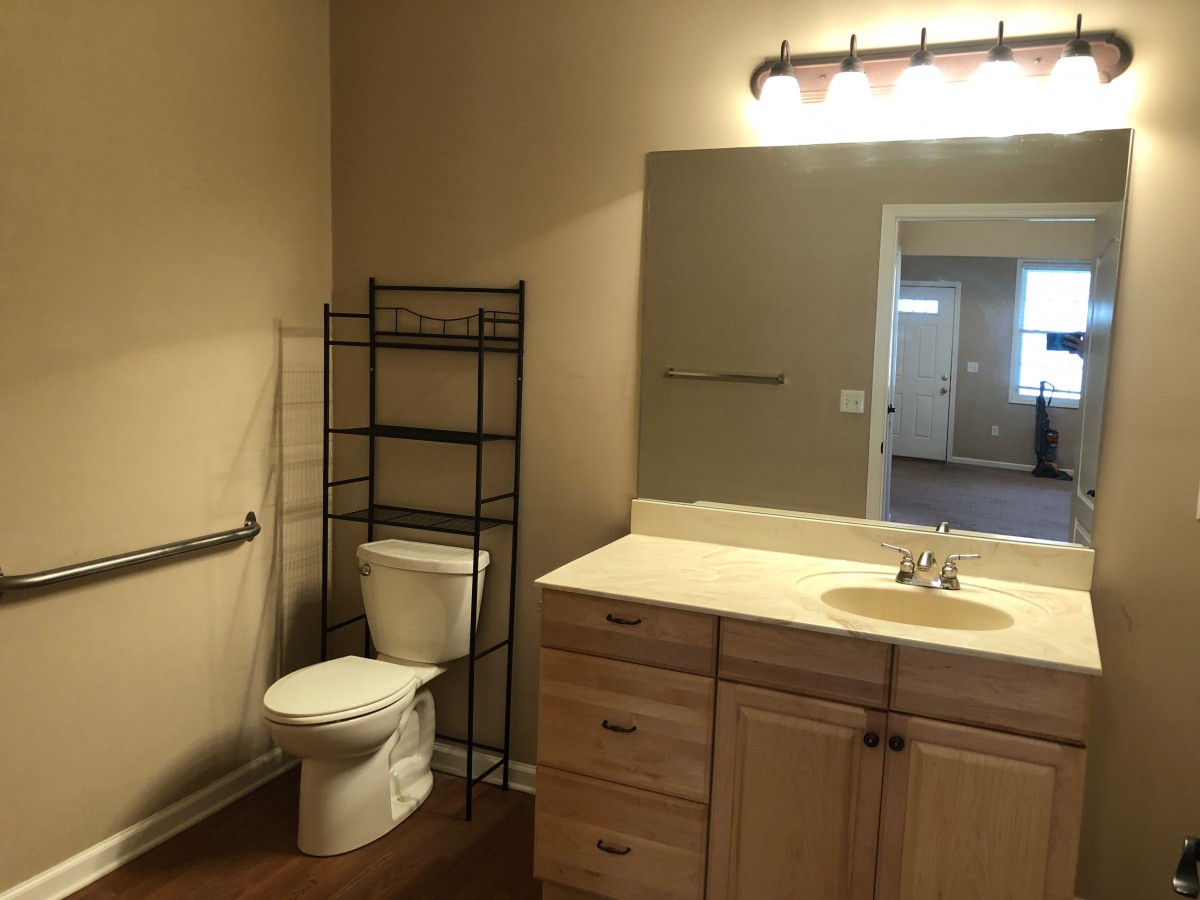 A bathroom with a toilet, sink, mirror, and storage shelf.