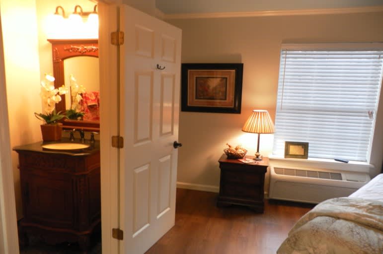 A cozy bedroom with a nightstand and a lamp next to the window, leading into an adjacent room.