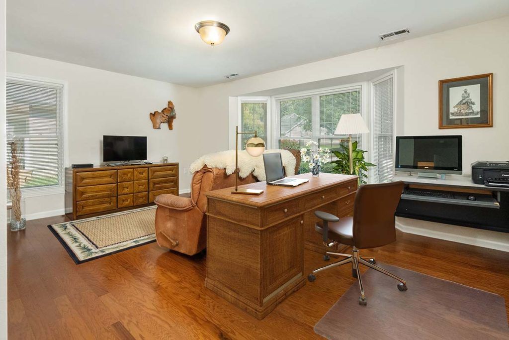 A cozy home office space with a wooden desk and chair, adjacent to a living area featuring a sofa, television, and large windows overlooking greenery.