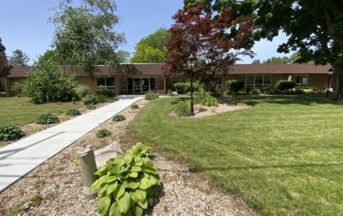 A landscaped area with a building in the background, featuring a sidewalk, trees, and well-maintained greenery.