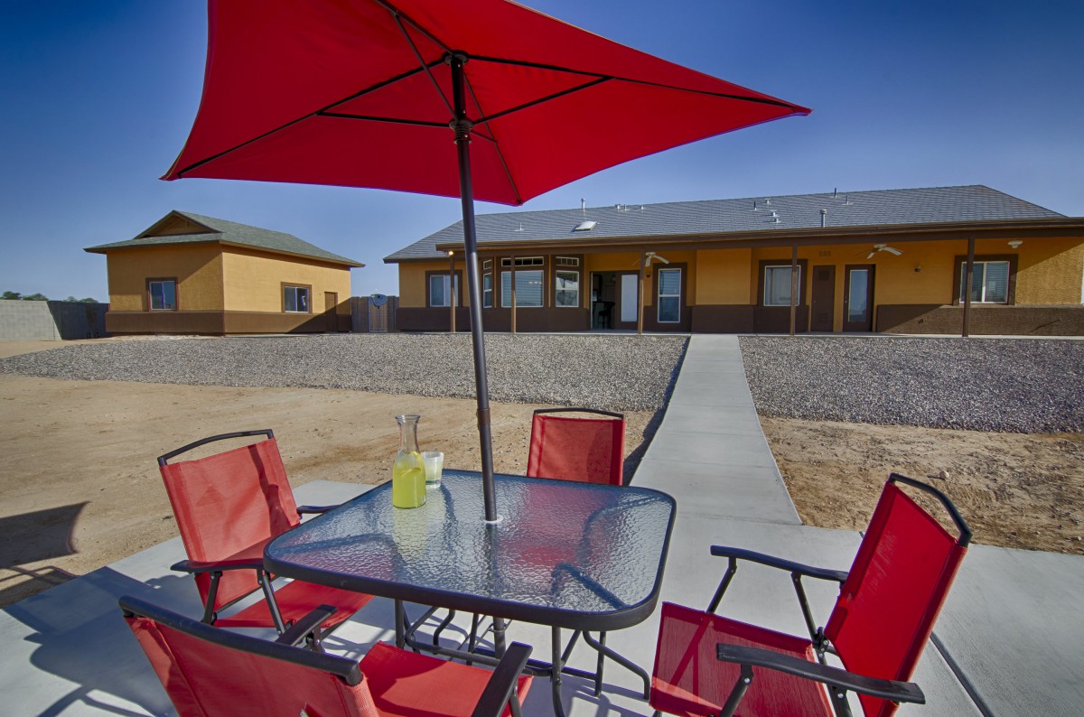 A patio area with a red umbrella and chairs set around a glass table, overlooking a house and backyard.