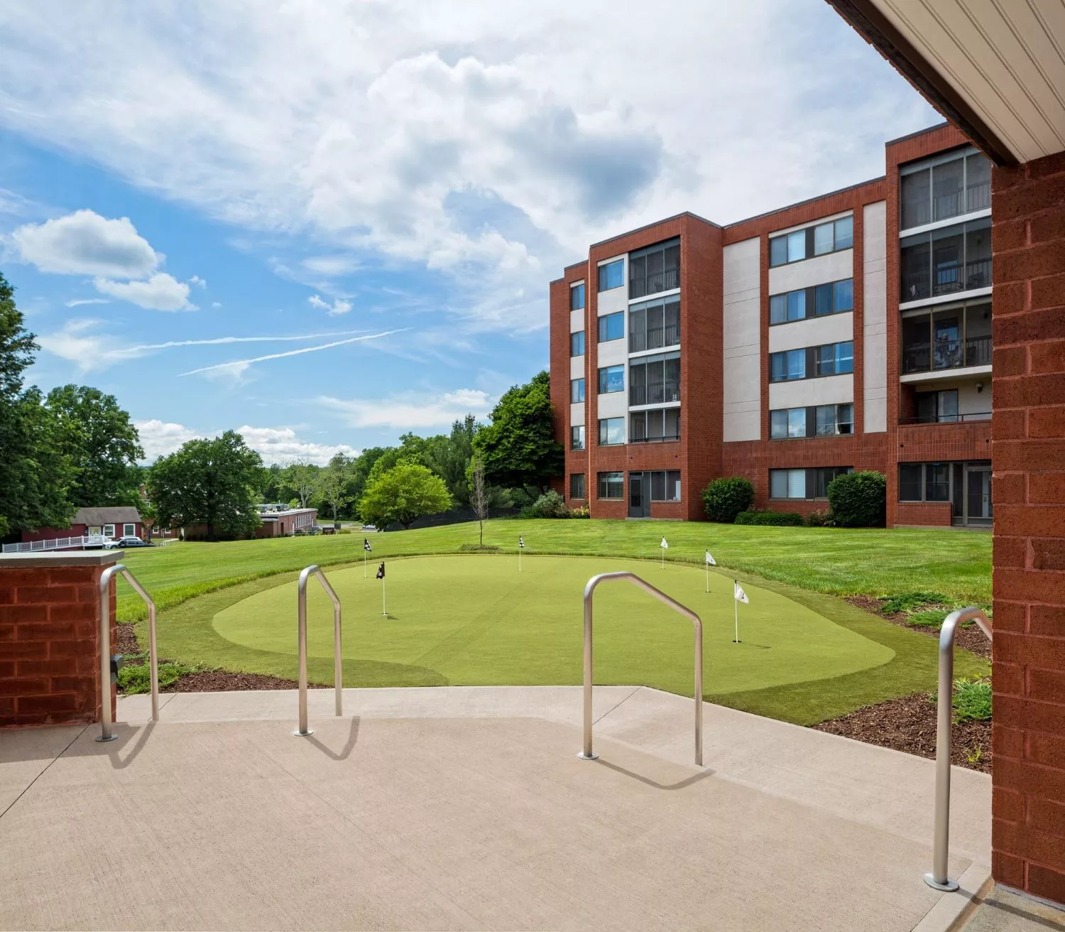 A view of a putting green surrounded by trees and a multi-story building under a partly cloudy sky.