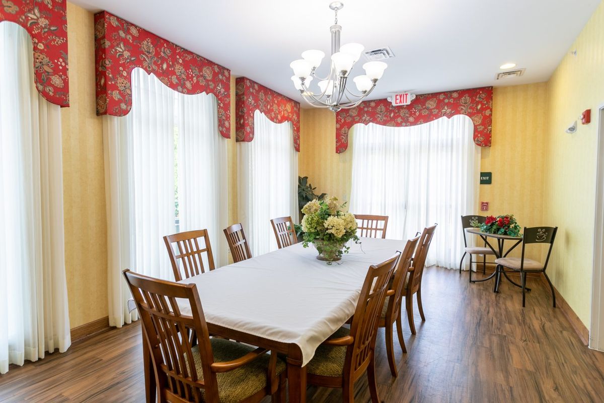 A well-lit dining room with a large table and wooden chairs, accented by red curtains and a chandelier overhead.
