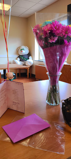 A beautifully arranged bouquet in a vase next to a heartfelt greeting card, with a large teddy bear in the background.
