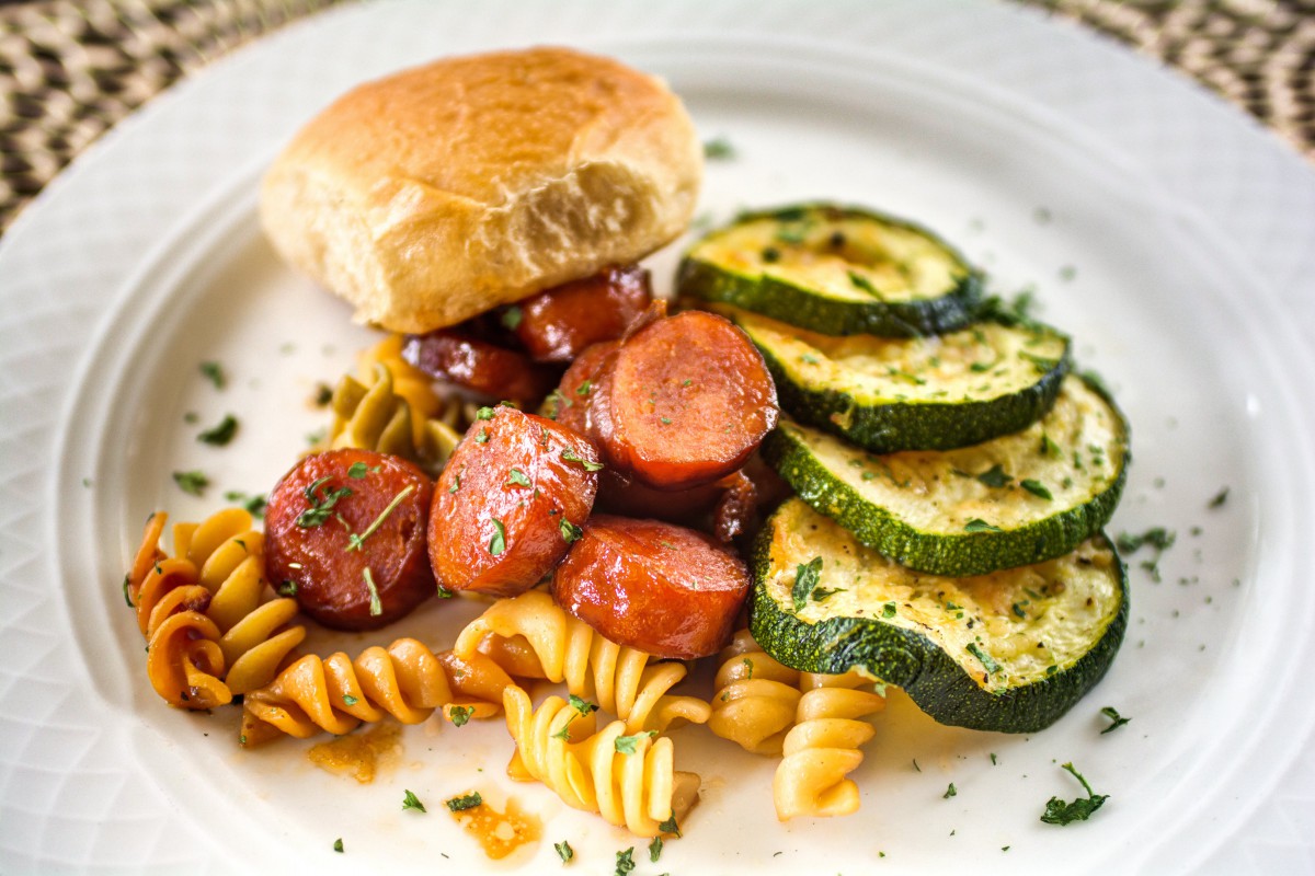 A delicious meal featuring sliced sausage, pasta, zucchini slices, and a bread roll on a white plate.